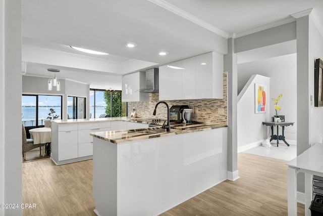 kitchen with wall chimney exhaust hood, decorative light fixtures, ornamental molding, kitchen peninsula, and white cabinets