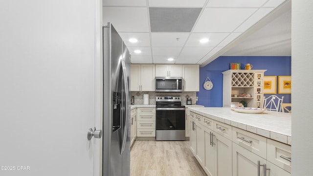 kitchen with tasteful backsplash, appliances with stainless steel finishes, light hardwood / wood-style flooring, and a drop ceiling