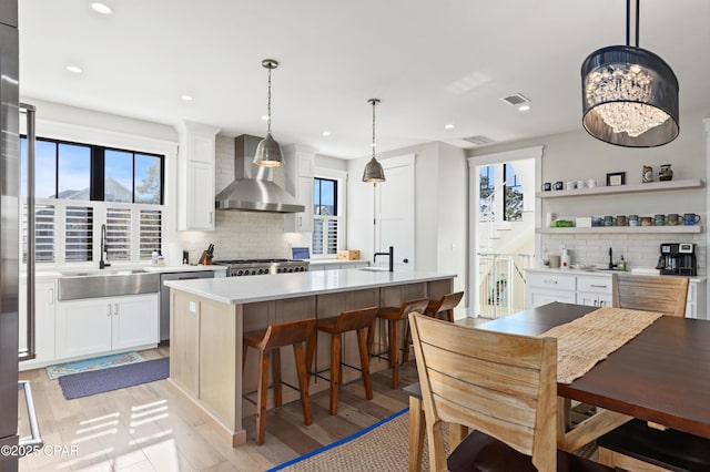 kitchen featuring wall chimney range hood, sink, white cabinetry, hanging light fixtures, and a kitchen island