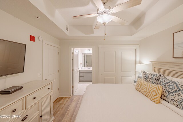 bedroom featuring ceiling fan, ensuite bathroom, a tray ceiling, and light hardwood / wood-style flooring