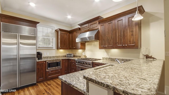 kitchen with a sink, a peninsula, light stone countertops, under cabinet range hood, and built in appliances