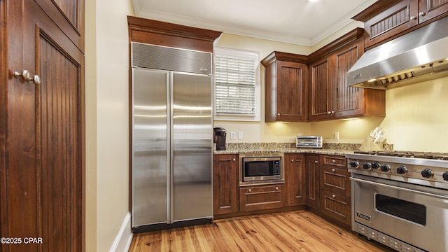 kitchen with built in appliances, under cabinet range hood, light wood-type flooring, light stone countertops, and crown molding