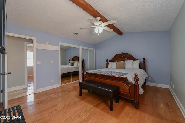 bedroom featuring ceiling fan, vaulted ceiling with beams, light hardwood / wood-style floors, a textured ceiling, and multiple closets