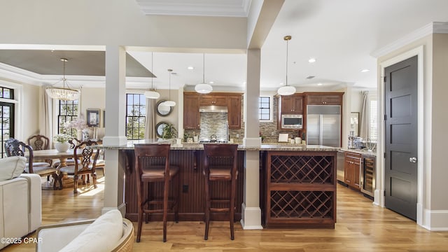 kitchen featuring decorative light fixtures, a breakfast bar area, beverage cooler, built in appliances, and light stone countertops