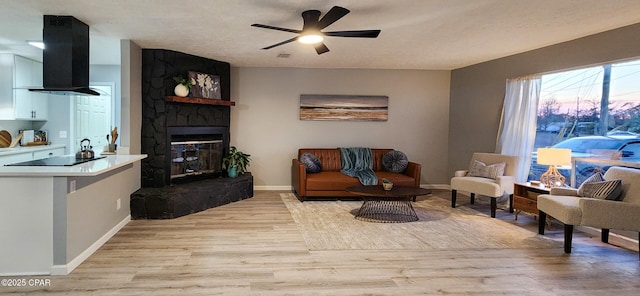 living room with ceiling fan, a textured ceiling, a stone fireplace, light wood-style flooring, and baseboards