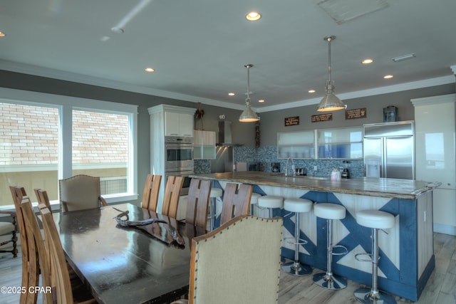 kitchen with wall chimney exhaust hood, white cabinetry, hanging light fixtures, kitchen peninsula, and built in fridge