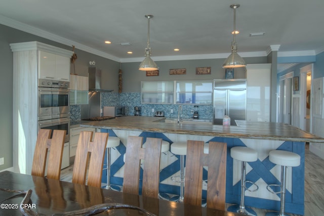 kitchen featuring wall chimney exhaust hood, stainless steel appliances, hanging light fixtures, and white cabinets