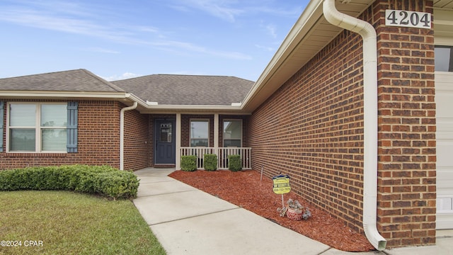 doorway to property featuring covered porch, brick siding, and roof with shingles