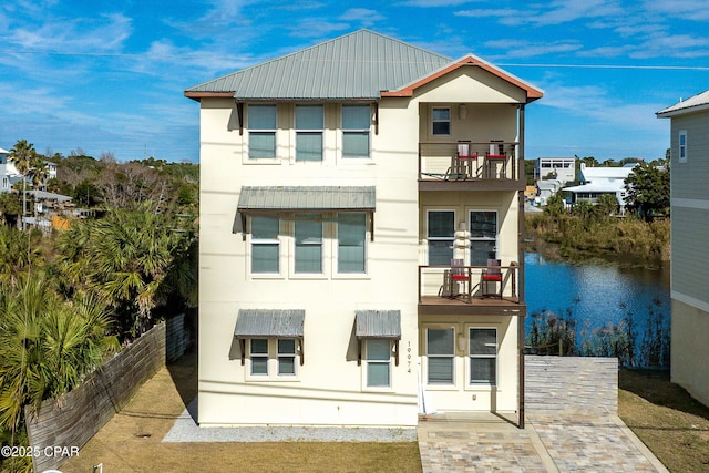 view of front of home featuring a balcony, metal roof, a water view, fence, and stucco siding