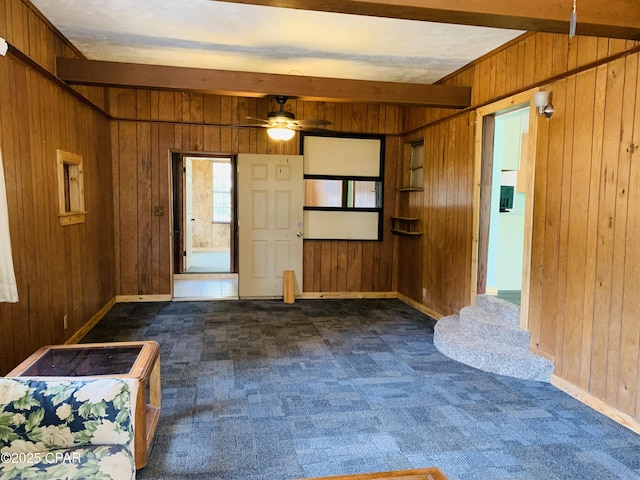 interior space with ceiling fan, wooden walls, baseboards, beam ceiling, and dark colored carpet