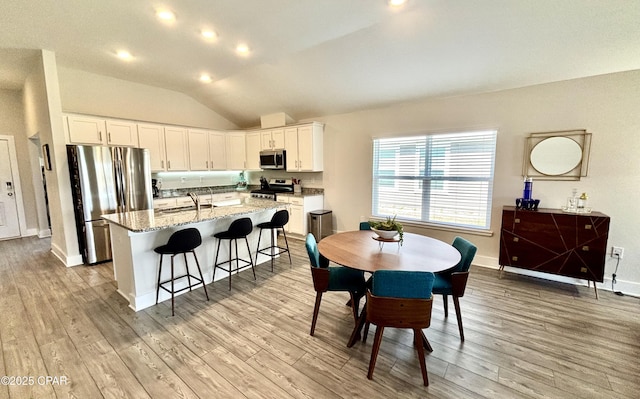 kitchen with light stone counters, a center island with sink, appliances with stainless steel finishes, vaulted ceiling, and light wood-type flooring