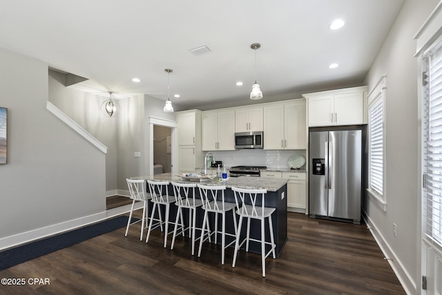 kitchen featuring appliances with stainless steel finishes, white cabinets, backsplash, hanging light fixtures, and a center island with sink