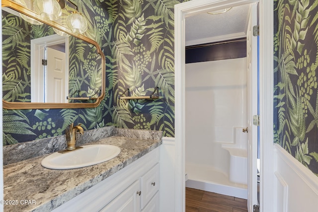 bathroom featuring a textured ceiling, wood finished floors, vanity, walk in shower, and wallpapered walls