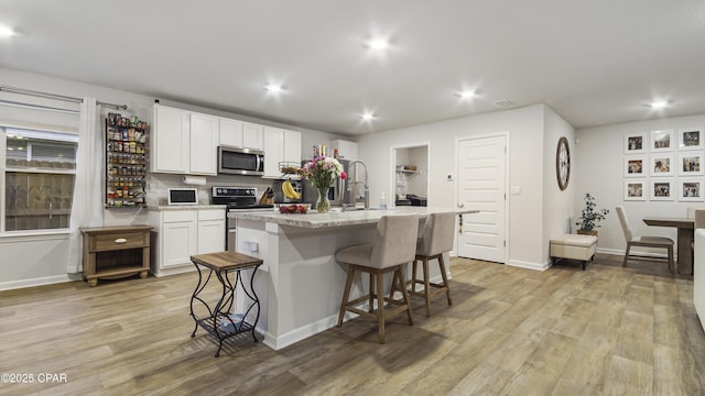 kitchen featuring appliances with stainless steel finishes, a kitchen island with sink, white cabinetry, light wood-type flooring, and a kitchen bar