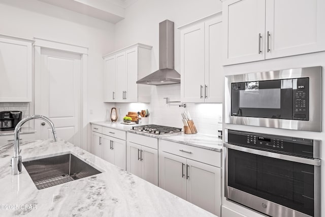 kitchen featuring stainless steel appliances, a sink, white cabinetry, wall chimney range hood, and light stone countertops