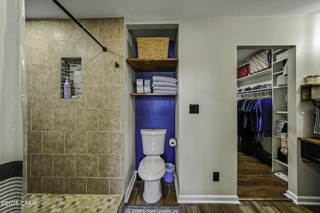 full bath with a walk in closet, toilet, a tile shower, wood finished floors, and baseboards