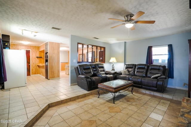 living area with a textured ceiling, light tile patterned floors, wood walls, visible vents, and a ceiling fan