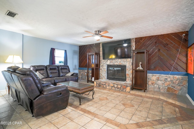 living room featuring visible vents, ceiling fan, a stone fireplace, wooden walls, and a textured ceiling