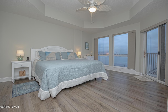 bedroom featuring access to exterior, a tray ceiling, a water view, wood finished floors, and baseboards