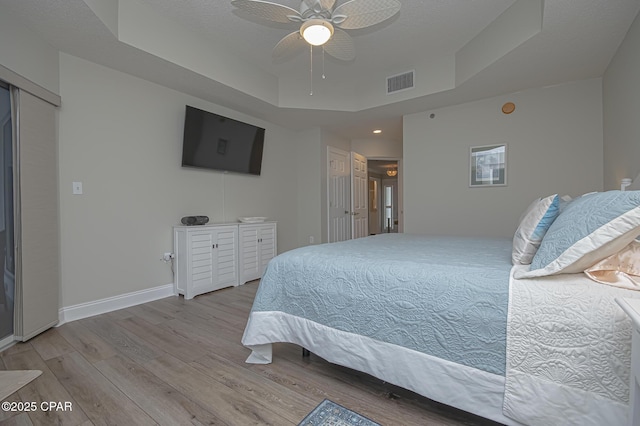 bedroom featuring ceiling fan, visible vents, baseboards, a tray ceiling, and light wood finished floors