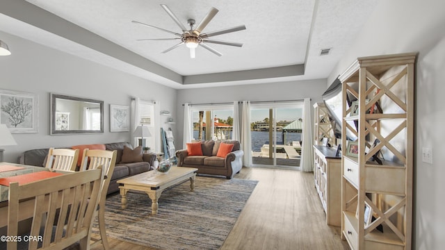 living area with a raised ceiling, visible vents, light wood-style floors, a ceiling fan, and a textured ceiling