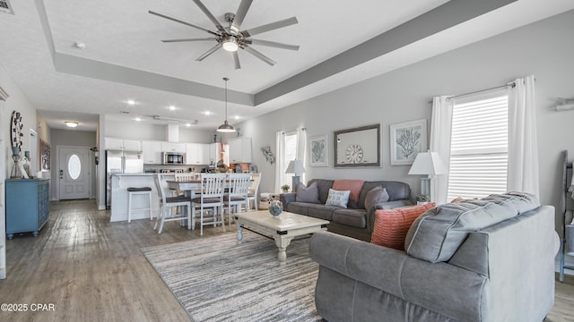 living room with a tray ceiling, dark wood-style flooring, a healthy amount of sunlight, and ceiling fan