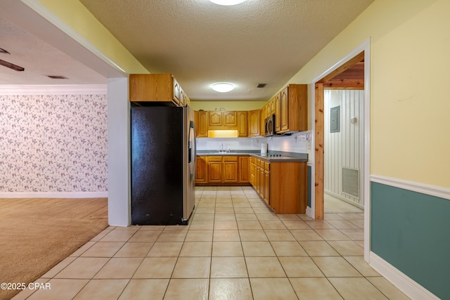 kitchen featuring a textured ceiling, light colored carpet, stainless steel appliances, brown cabinets, and wallpapered walls