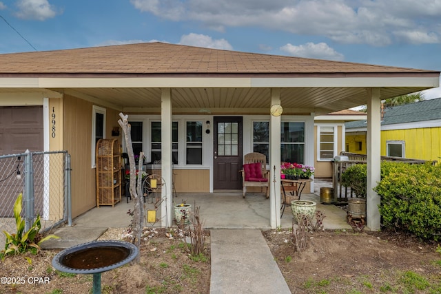 view of exterior entry with covered porch, a shingled roof, and fence