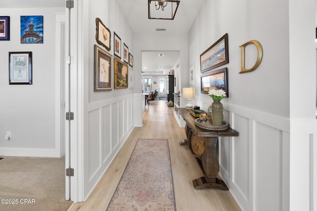 hall with light wood-style floors, visible vents, a decorative wall, and wainscoting