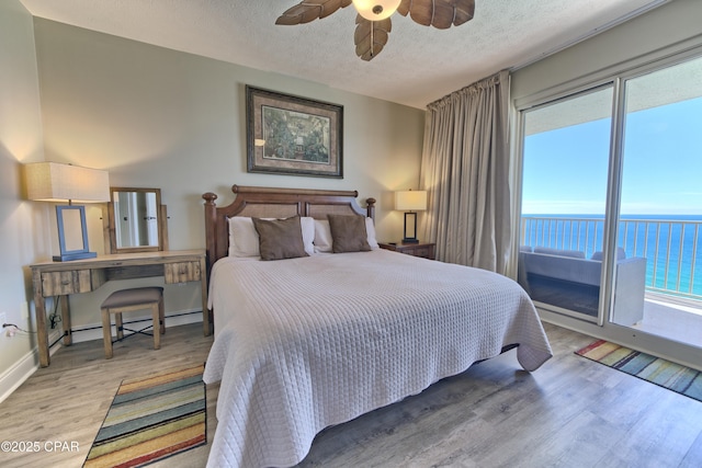 bedroom with wood finished floors, a water view, access to outside, a textured ceiling, and a baseboard heating unit