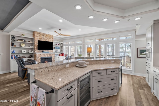 kitchen featuring beverage cooler, oven, a raised ceiling, a stone fireplace, and a ceiling fan