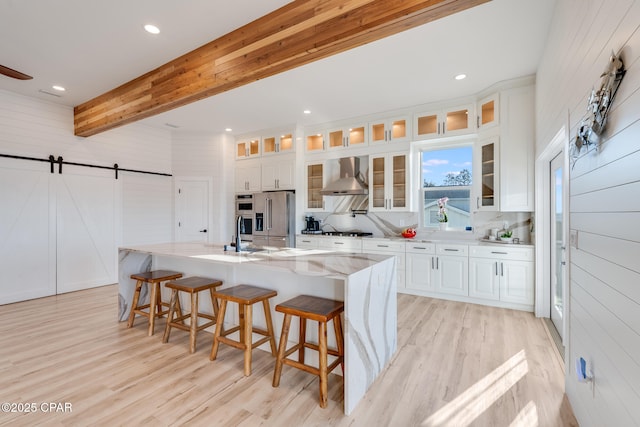 kitchen with a barn door, high end refrigerator, wall chimney range hood, gas stovetop, and beam ceiling