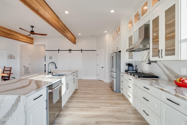 kitchen with beam ceiling, a barn door, appliances with stainless steel finishes, a sink, and wall chimney range hood