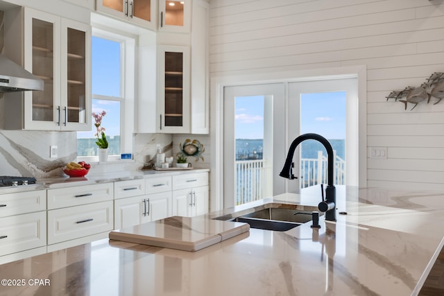 kitchen with light stone counters, a sink, white cabinets, wall chimney range hood, and tasteful backsplash