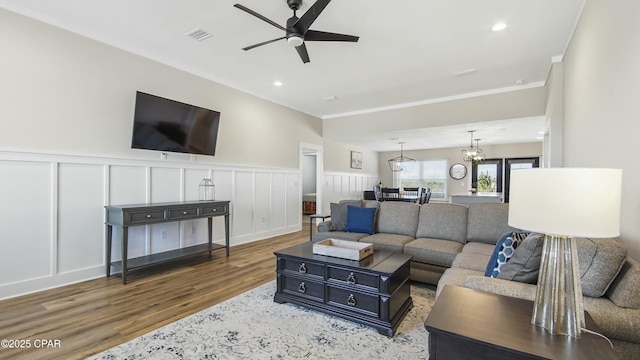 living room featuring crown molding, visible vents, wainscoting, wood finished floors, and ceiling fan with notable chandelier