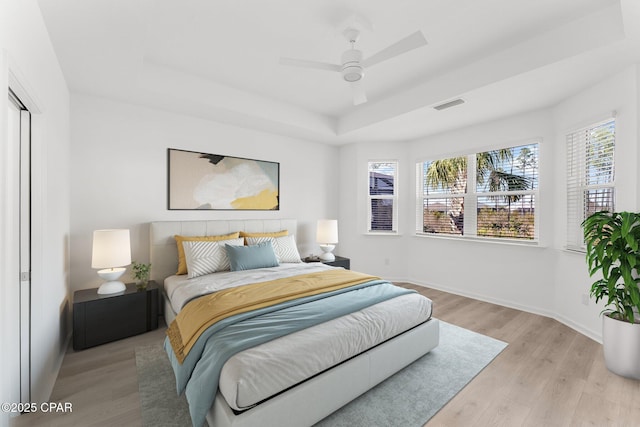 bedroom featuring visible vents, baseboards, light wood finished floors, a raised ceiling, and ceiling fan