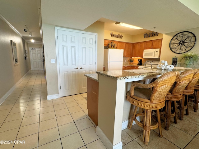 kitchen with brown cabinetry, light tile patterned flooring, light stone countertops, white appliances, and a peninsula