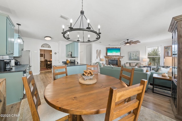 dining room featuring visible vents, crown molding, ceiling fan with notable chandelier, light wood-style floors, and a glass covered fireplace