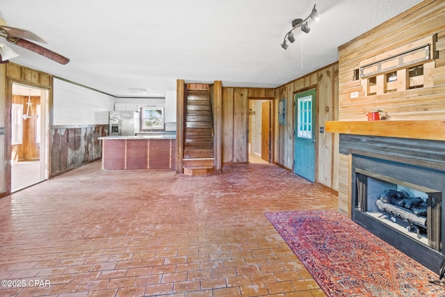 unfurnished living room with a textured ceiling, brick floor, wooden walls, a fireplace, and a ceiling fan