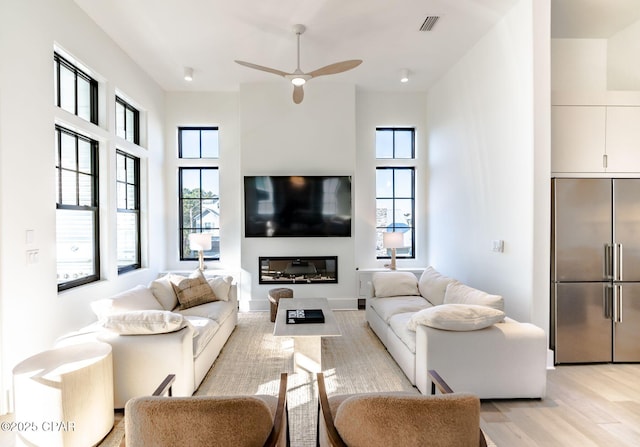 living area with a ceiling fan, light wood-type flooring, visible vents, and a glass covered fireplace