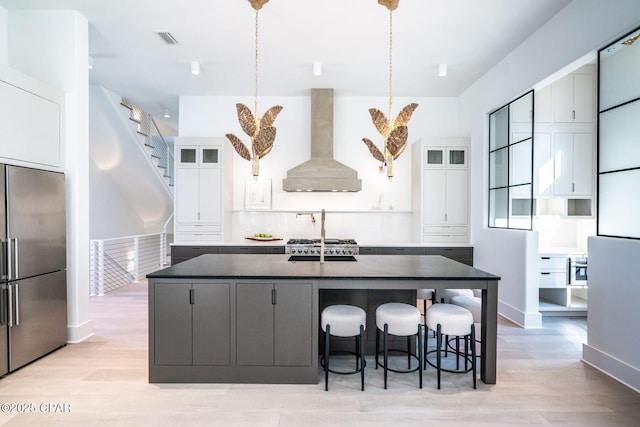 kitchen featuring a breakfast bar, wall chimney range hood, an island with sink, light wood-type flooring, and stainless steel fridge