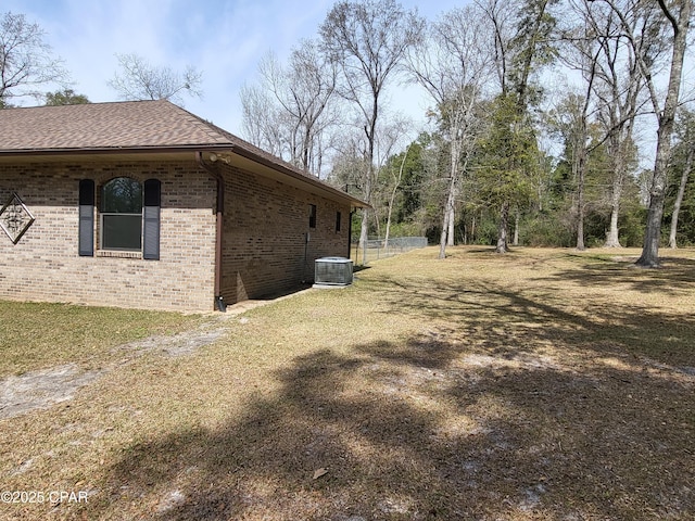 view of side of home with brick siding, roof with shingles, a lawn, fence, and cooling unit