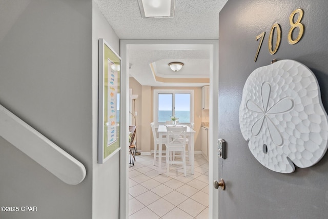 hall featuring a raised ceiling, a textured ceiling, baseboards, and light tile patterned floors