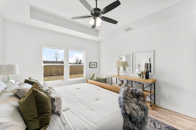 bedroom with a tray ceiling, a ceiling fan, visible vents, wood finished floors, and baseboards