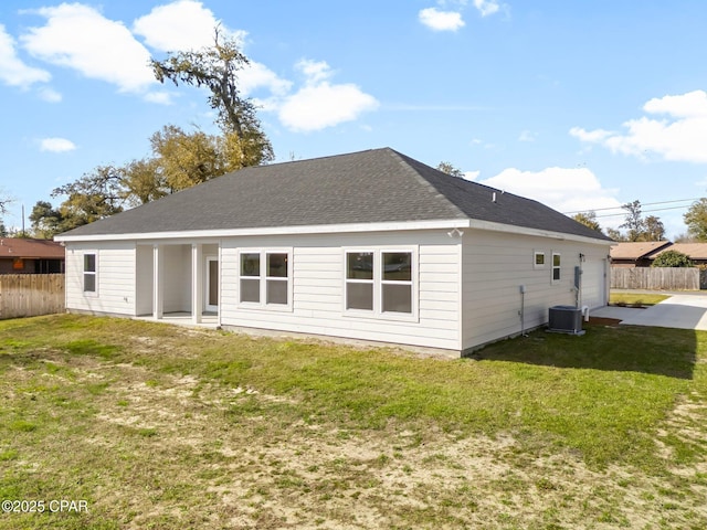 rear view of house with central AC unit, a shingled roof, fence, and a lawn