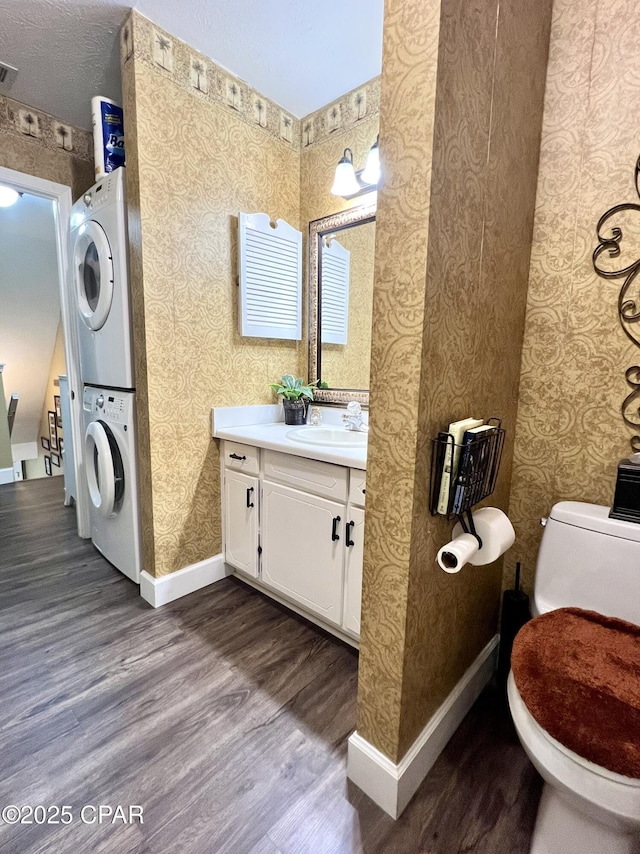 bathroom featuring stacked washer / drying machine, toilet, wood finished floors, baseboards, and wallpapered walls