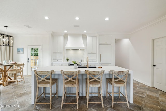 kitchen featuring premium range hood, a kitchen island with sink, ornamental molding, and light countertops