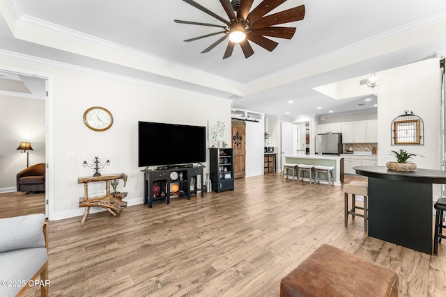 living area featuring a barn door, baseboards, light wood-style floors, ornamental molding, and a raised ceiling