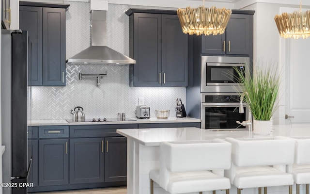 kitchen with light countertops, backsplash, appliances with stainless steel finishes, a chandelier, and wall chimney exhaust hood