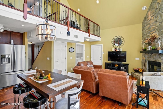 living area with visible vents, a towering ceiling, wood finished floors, a stone fireplace, and ceiling fan with notable chandelier